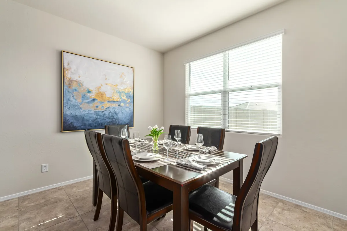 A bright, inviting dining area is shown with a family-style dining table, modern artwork on the wall, and a window flooding the space with natural light. The scene suggests lively meals and shared moments.