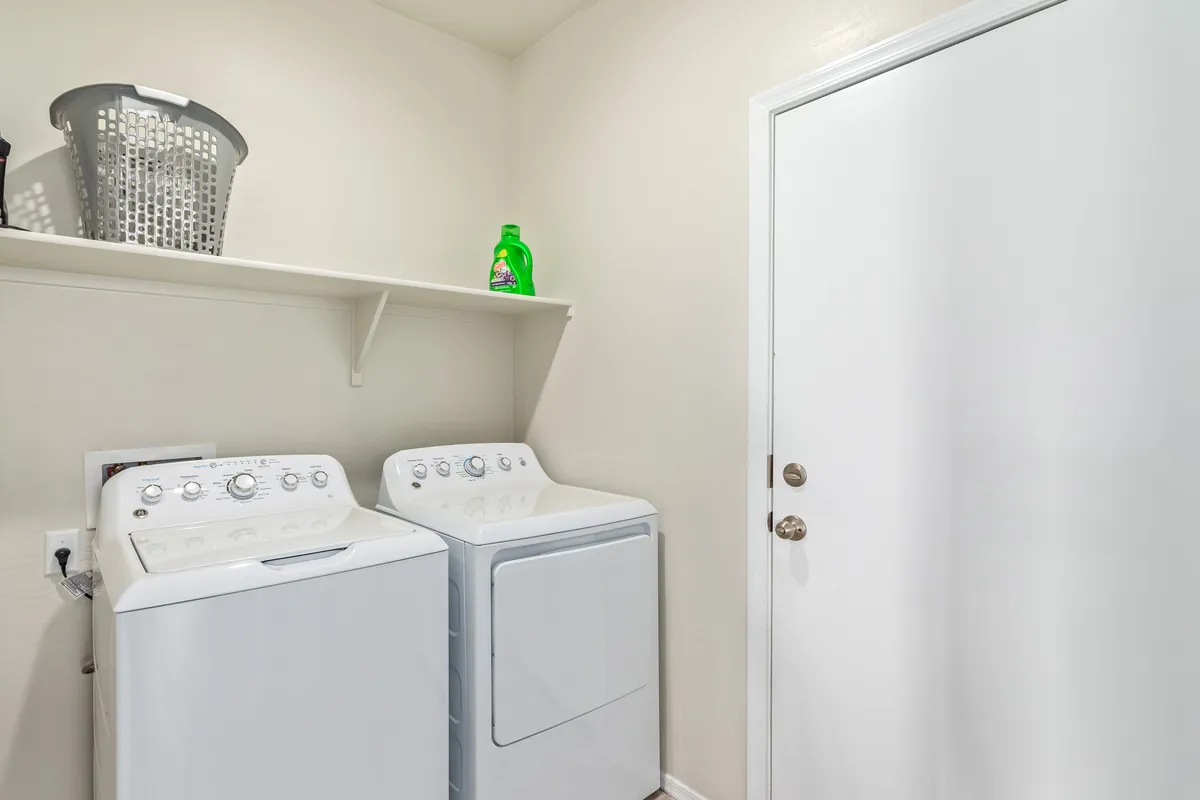 A tidy laundry room featuring a modern washing machine and dryer, a clean white shelf above with detergents, and a basket ready for use. The space is well-organized, emphasizing functionality and convenience for daily chores.