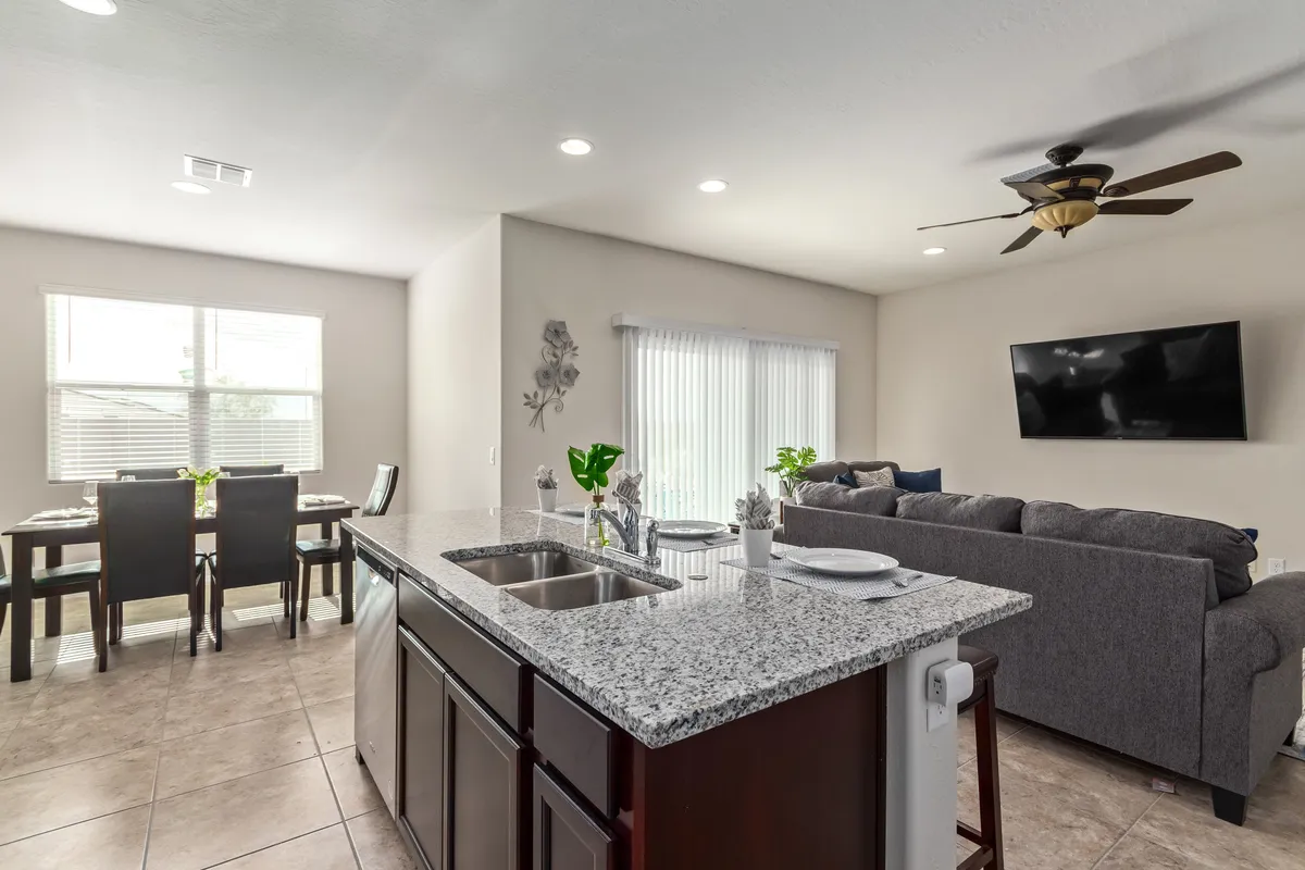 This image captures the open-concept great room, prominently featuring the granite kitchen island, a large dining table, a comfortable sofa, and a wall-mounted TV. The kitchen is visible and equipped, while the living area offers plush seating that invites relaxation and socializing. The space is brightly lit and arranged for both cooking and entertaining.