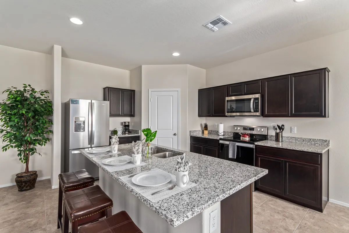 A warm, functional kitchen space with granite countertops, stainless steel appliances, bar stools, and a houseplant. The kitchen is modern yet inviting, clearly suited for preparing meals and hosting family or guests. Natural light and the accessible setup evoke a sense of returning home to a welcoming, well-equipped environment.