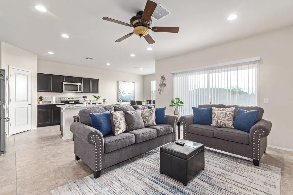 A cozy living area featuring plush sofas, a coffee table, a ceiling fan overhead, and a clear view of the connected kitchen area. The setting is ideal for relaxing, entertaining guests, or enjoying a quiet evening. The layout demonstrates the seamless flow between living and kitchen spaces.